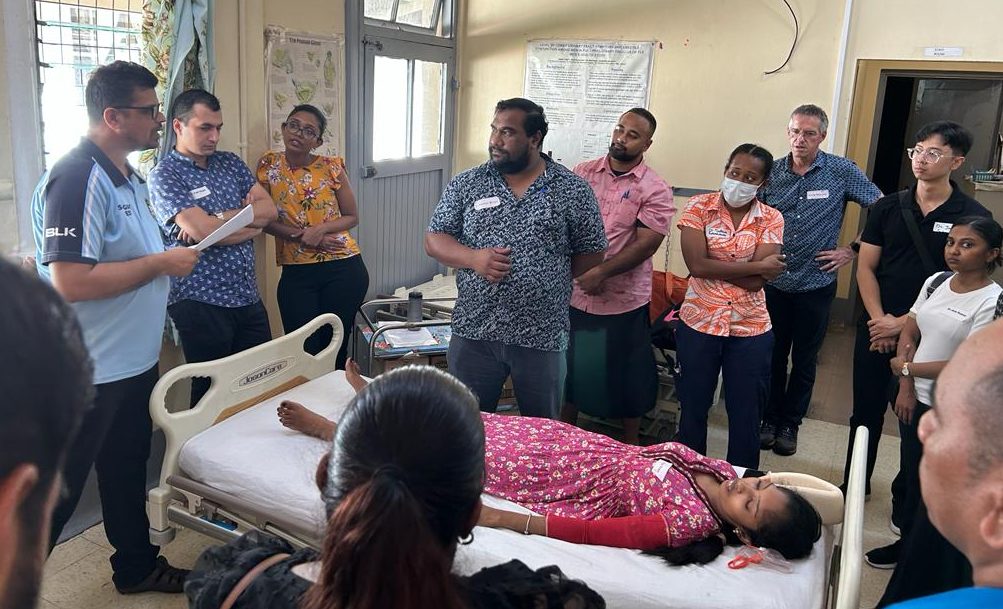In a training workshop, a group of people gather around a woman posing in a hospital bed, demonstrating management of a burn injury.