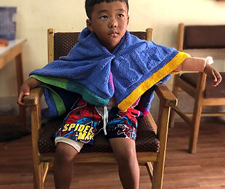Kinley, a young boy, sitting up in a chair with a blanket pinned around his shoulders like a cape.