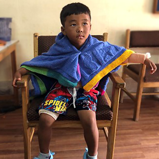 Kinley, a young boy, sitting up in a chair with a blanket pinned around his shoulders like a cape.