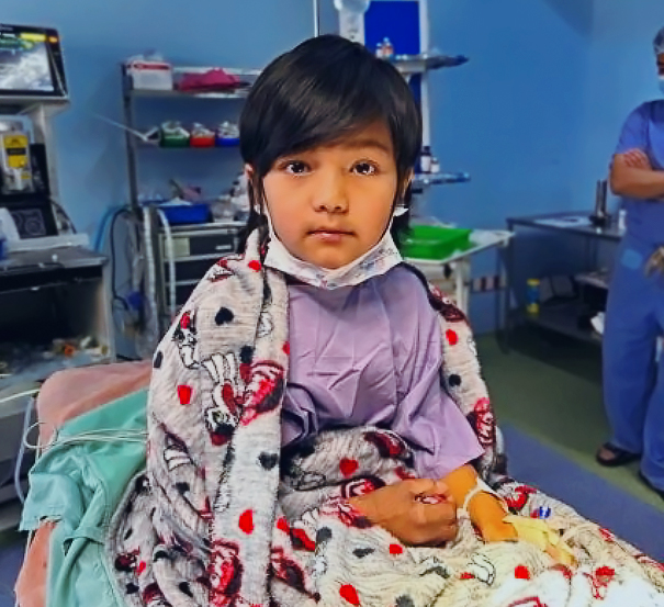 A young girl in a hospital gown sits up on a bed with a blanket around her shoulders and hands in her lap. She looks at the camera.