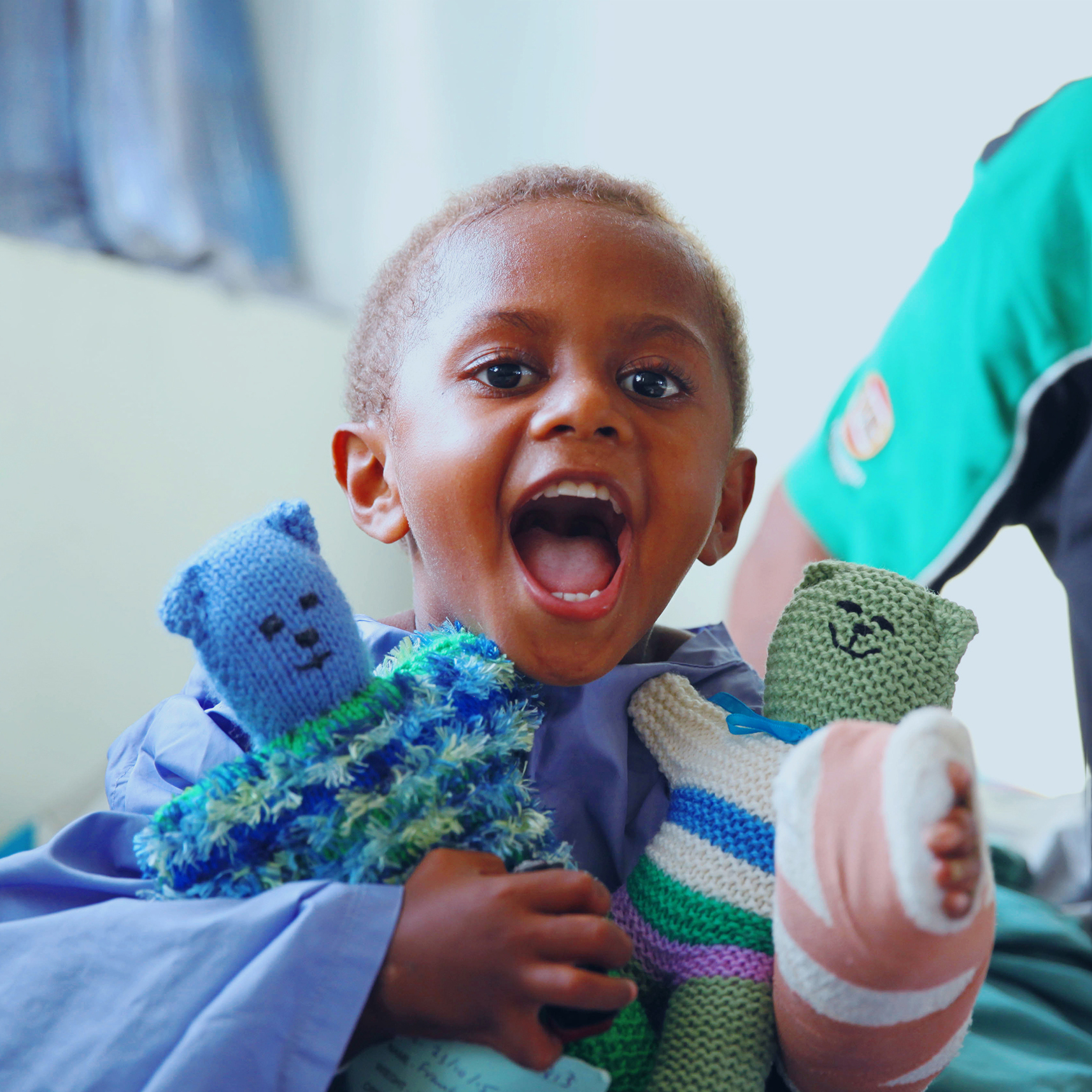 Young boy with a bandaged hand hugs two teddies and smiles excitedly.