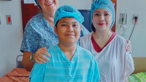 A young boy in a hospital gown smiles for the camera with his mum and another woman dressed in scrubs.