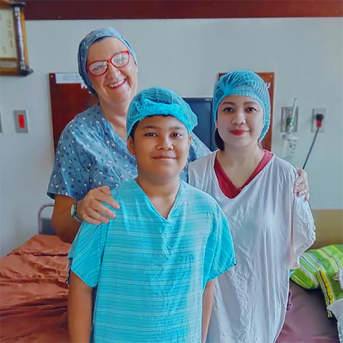 A young boy in a hospital gown smiles for the camera with his mum and another woman dressed in scrubs.