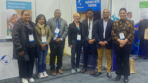 A group of seven people with matching lanyards stand in front of the Interplast booth in a conference hall.