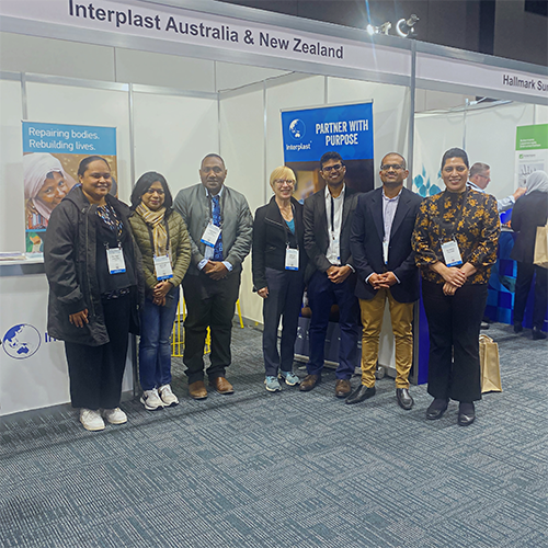 A group of seven people with matching lanyards stand in front of the Interplast booth in a conference hall.