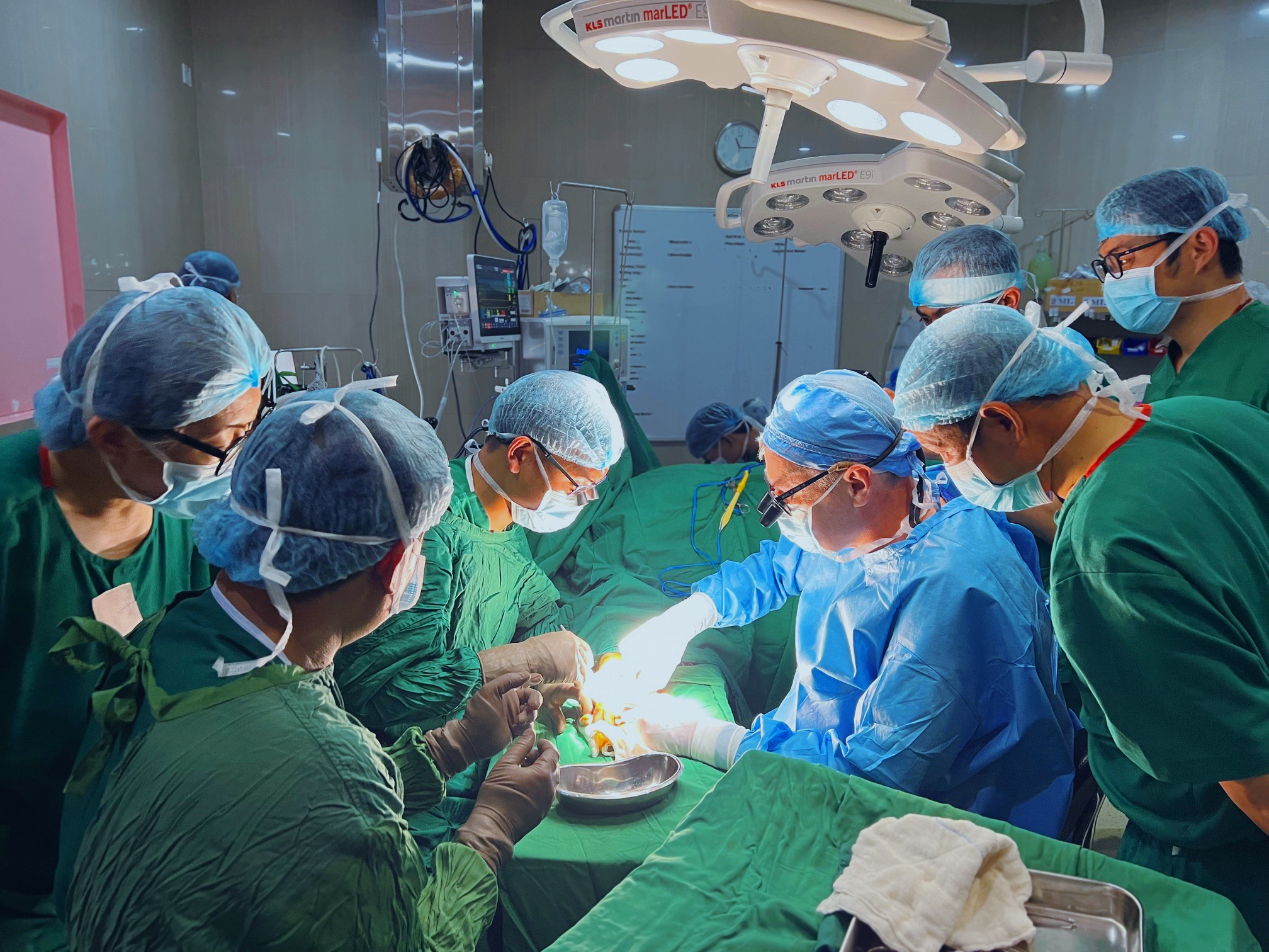 Two surgeons are supported by a team of at least 5 other people to operate on an unseen patient.