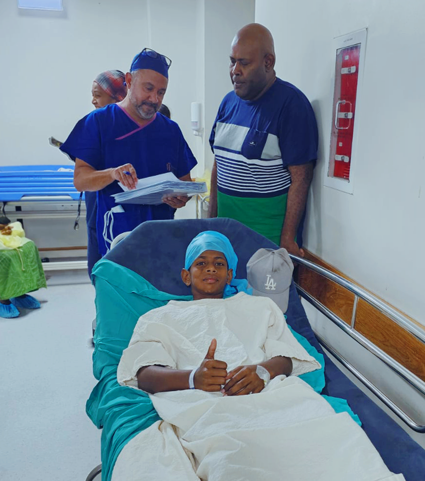 A young patient in a surgical gown gives a thumbs up from their hospital bed.