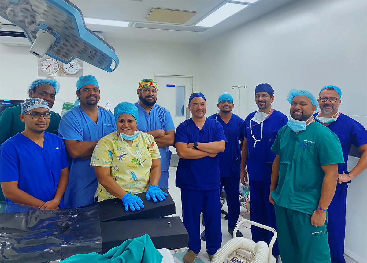 10 people in surgical scrubs stand in a clean operating theatre and smile at the camera.