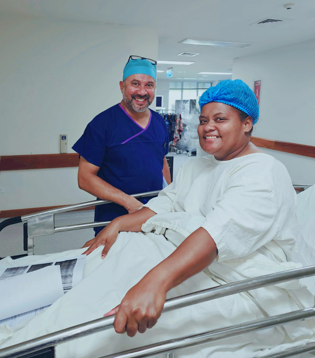 A person in a hospital gown sits up in their hospital bed. She smiles for a photo with a staff member in surgical scrubs.