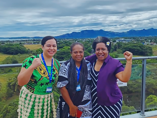 Three women smile and pose triumphantly in front of the Fiji landscape.