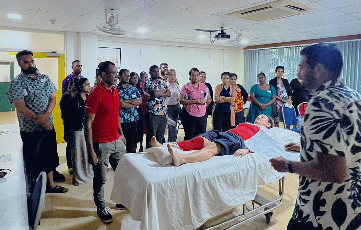 Inside a classroom, a group gathers around a hospital bed where a person with limbs and torso painted red lies.