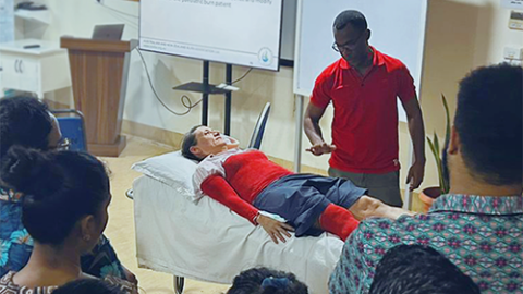 Inside a classroom, a person with limbs and torso painted red lies on a bed while an instructor speaks to a group of people about them.