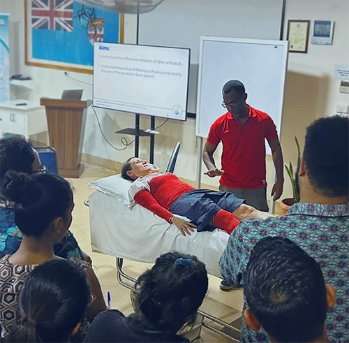 Inside a classroom, a person with limbs and torso painted red lies on a bed while an instructor speaks to a group of people about them.