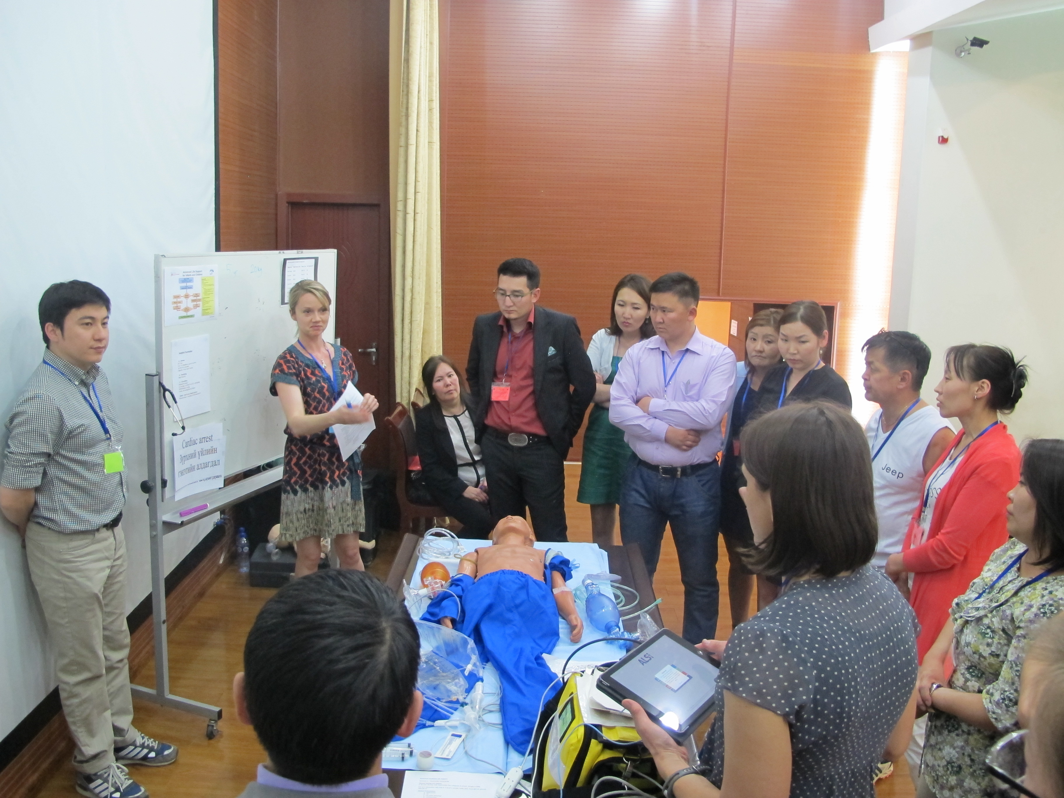 A large group gather  around a model on a table and whiteboard in discussion.