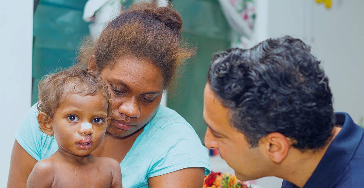 A young child with a bandage on their nose is held by their mother while an Interplast volunteer talks to them softly.