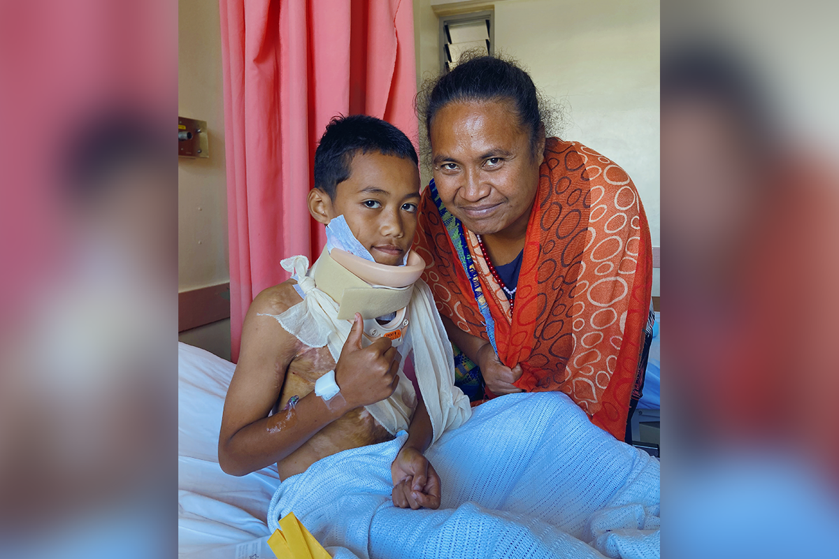 A young boy in a neck brace and with upper torso bandaged sits up in his hospital bed and smiles for a photo with his mother. He smiles gives a thumbs up.