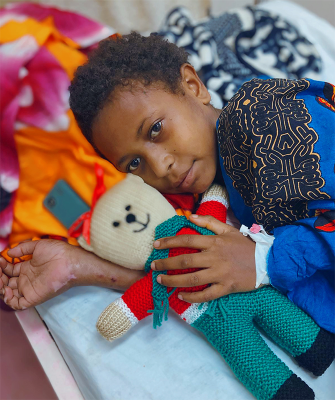 A young boy lies on his side in bed, holding a teddy bear with a bandaged hand.