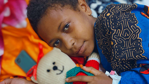 A young boy lies on his side in bed, holding a teddy bear with a bandaged hand.