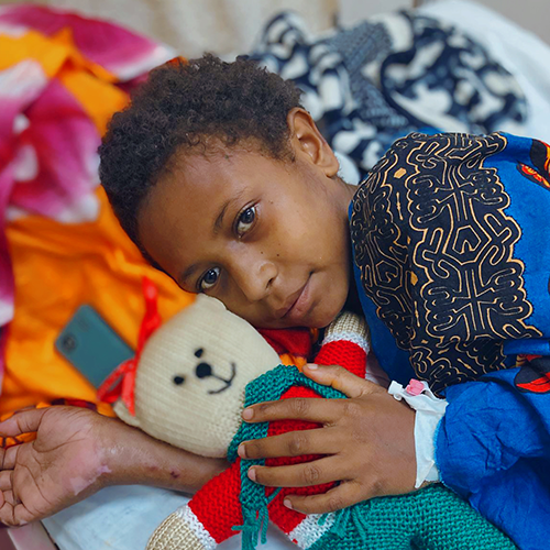 A young boy lies on his side in bed, holding a teddy bear with a bandaged hand.