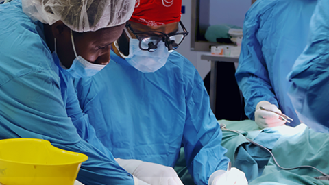 Two people in surgical scrubs delicately perform surgery on the hand of an unseen patient.