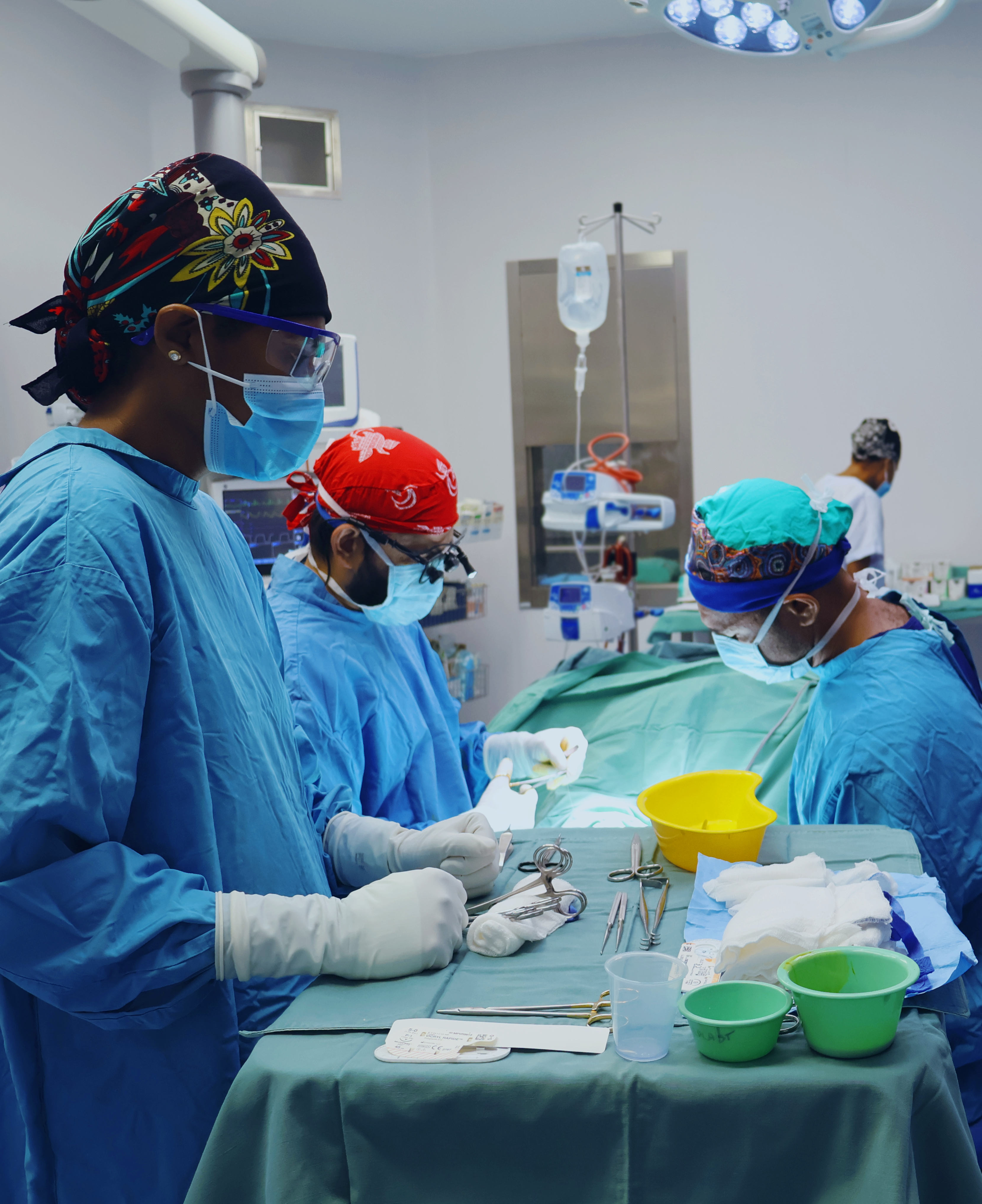 A nurse in surgical scrubs keeps track of all equipment while two surgeons work on an unseen patient.