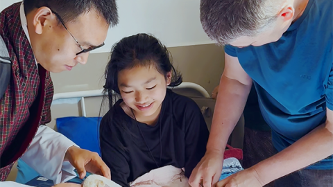 Two men take care to undress the bandages on the hands of a young girl, who watches them smiling.