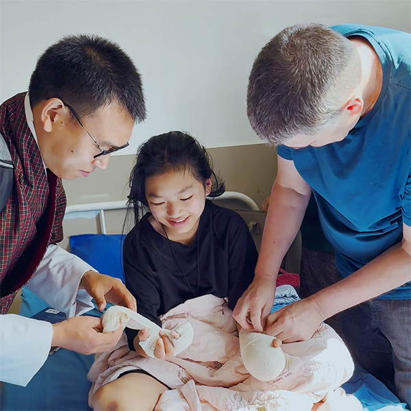 Two men take care to undress the bandages on the hands of a young girl, who watches them smiling.