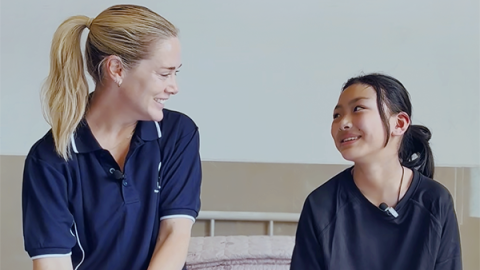 A young girl with dressings on her hands and a young woman sit together on a hospital bed, smiling at each other.