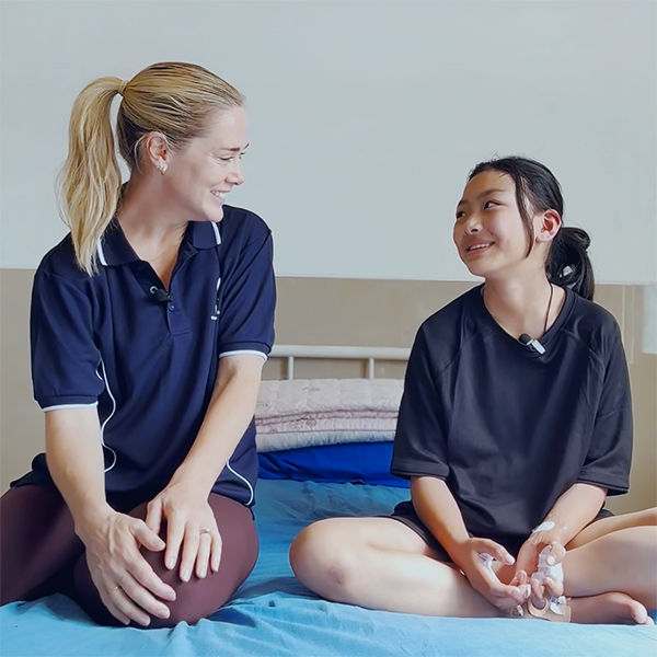 A young girl with dressings on her hands and a young woman sit together on a hospital bed, smiling at each other.