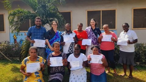 A group of eleven people hold certificates and pose for a photo in front of a building. Seven of them stand while four of them kneel on the grass below.