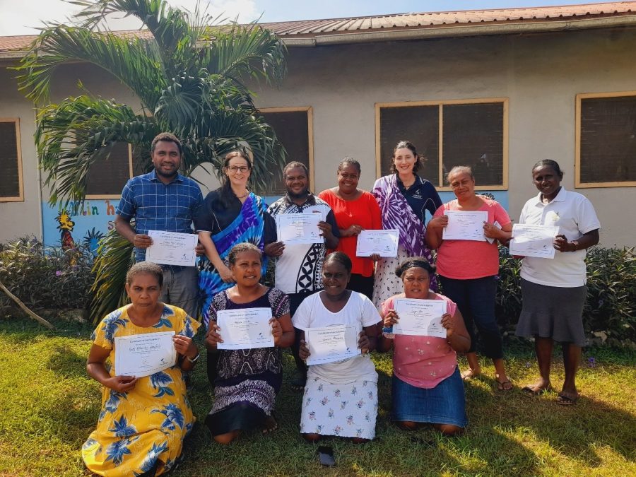 A group of eleven people hold certificates and pose for a photo in front of a building. Seven of them stand while four of them kneel on the grass below.